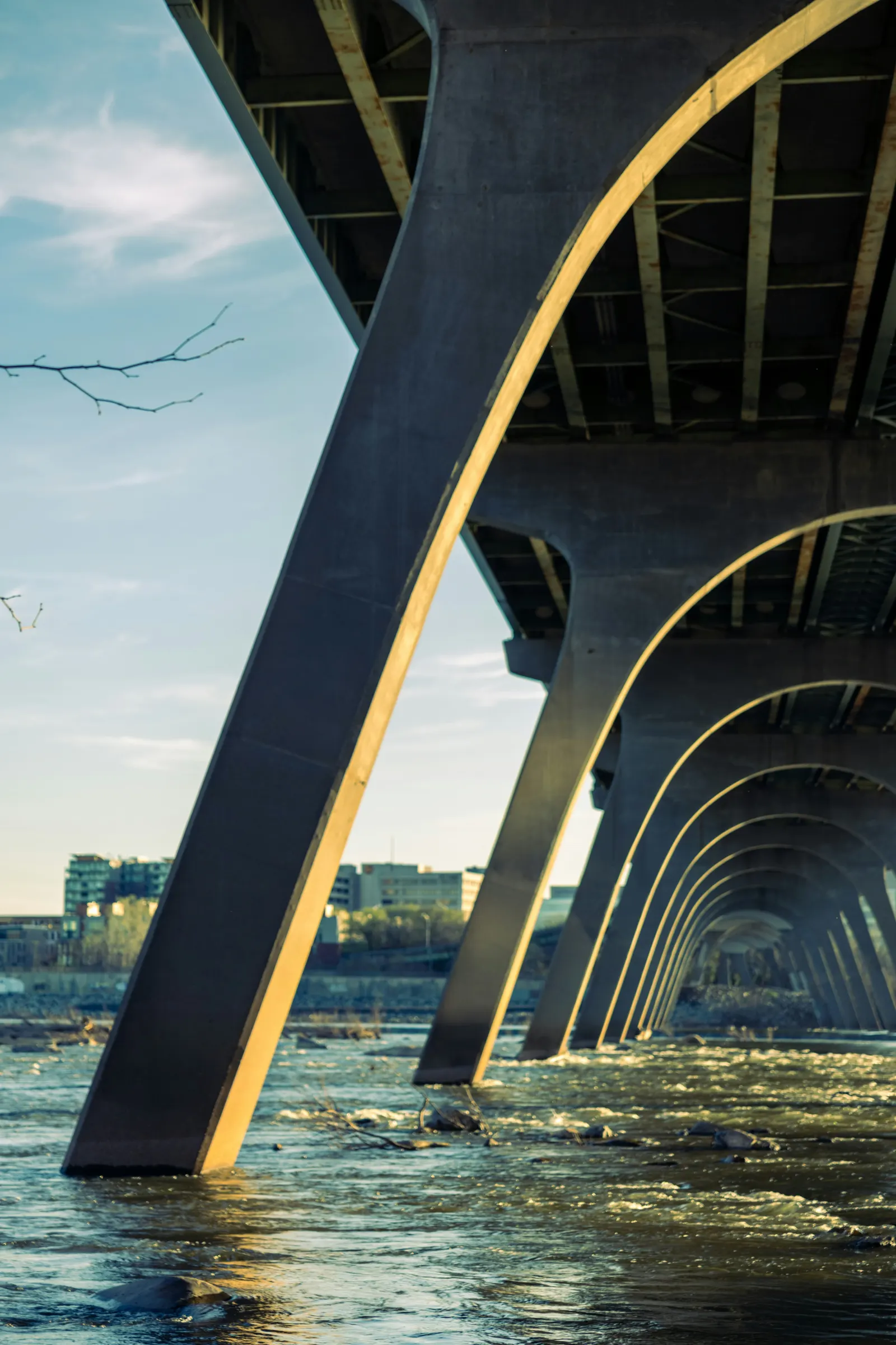 Sunlit concrete arches of a bridge receding over a rocky river, with the Richmond, Virginia skyline beyond.